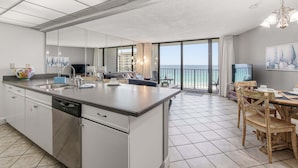 A modern kitchen with a large island, a dining area with wooden chairs, and a balcony with a view of the sea. - Hello Sunshine Edgewater, Tower 1, #905-Beachfront! 2Bd-2Ba-Sleeps 6-Fun In The Sun! (Panama City Beach)