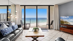 A modern living room with a sofa, coffee table, and TV, overlooking a beach. - Hello Sunshine Edgewater, Tower 1, #905-Beachfront! 2Bd-2Ba-Sleeps 6-Fun In The Sun! (Panama City Beach)