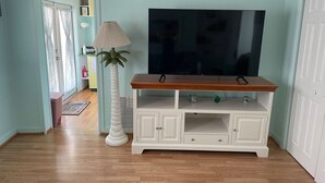 Una sala de estar con piso de madera, un mueble blanco para entretenimiento con un televisor y una lámpara alta de color blanco.