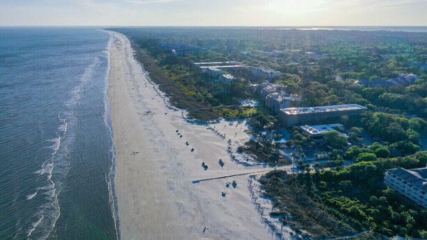 An aerial view of a beach with a long stretch of sand, a few buildings, and dense greenery. - Balcony overlooks BEAUTIFUL OCEAN VIEW, just steps away from sand! (Hilton Head Island)