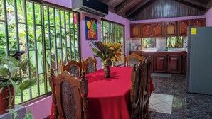 A dining area with a red table, wooden chairs, and a large window with a view of greenery. - Ceibo Arenal Home (La Fortuna)