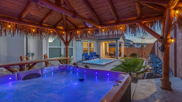 Ein Whirlpoolbereich mit Holzbalken und Lichterketten, umgeben von Gartenmöbeln und mit Blick auf ein Haus im Hintergrund.
