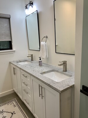 Une salle de bain avec deux lavabos, une fenêtre avec des stores, un porte-serviettes et un miroir.