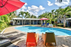 Ein Pool mit Liegestühlen und Sonnenschirmen, umgeben von Palmen und einem überdachten Bereich mit Sitzgelegenheiten.
