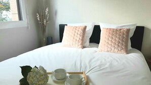 Cama bem arrumada com roupa de cama branca e duas almofadas decorativas, bandeja com duas chávenas e um arranjo floral, e uma janela com vista para árvores.