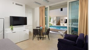 Ein modernes Hotelzimmer mit Flachbildfernseher, einer Essecke mit Glastisch und Stühlen, einer Couch und einer Schiebetür, die auf einen Balkon mit Pool führt.