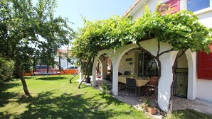 Un espace extérieur abrité, aménagé pour prendre les repas, avec une table et des chaises, entouré de piliers blancs et d’un arbre aux feuilles vertes.