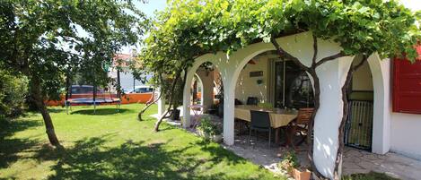 A covered outdoor dining area with a table and chairs, surrounded by white pillars and a tree with green leaves.