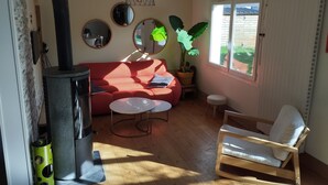 Un salon chaleureux avec un canapé rouge, une table basse ronde, une chaise en bois, un radiateur noir et une grande fenêtre donnant sur l’extérieur.