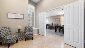 Un pasillo espacioso con pared beige, una cita enmarcada, dos sillones estampados y una puerta blanca que da a un comedor.