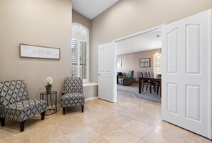 A spacious hallway with a beige wall, a framed quote, two patterned chairs, and a white door leading to a dining area.
