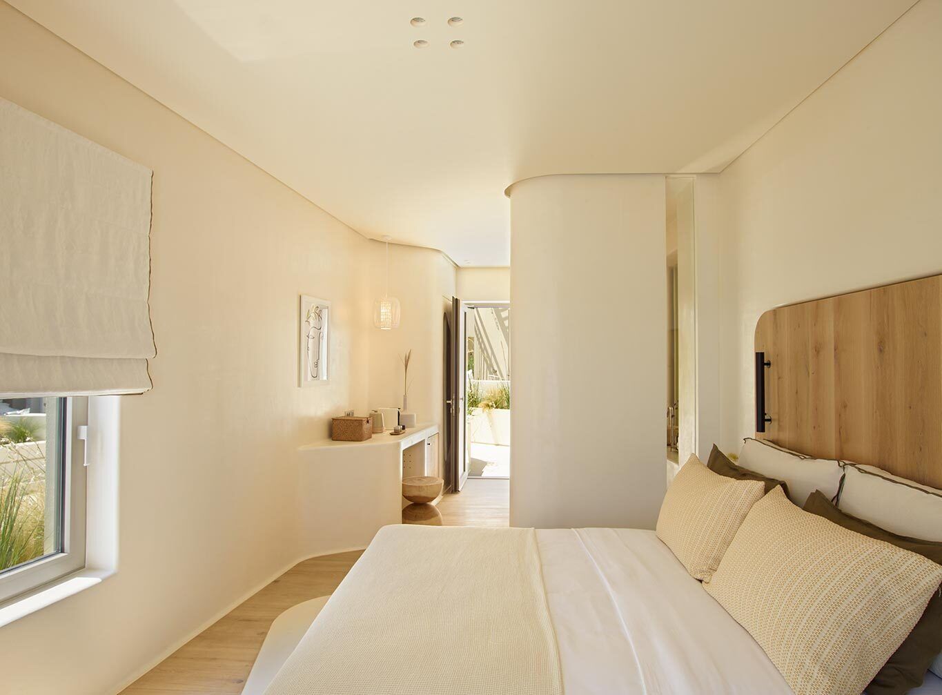 A modern bedroom with a large bed, a wooden headboard, a bedside table with a lamp, and a view of the outdoors.