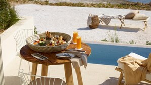 A dining table with a basket of food and drinks, a poolside chair, and a lounge chair with a towel. - White Pebble Suites (Milos)