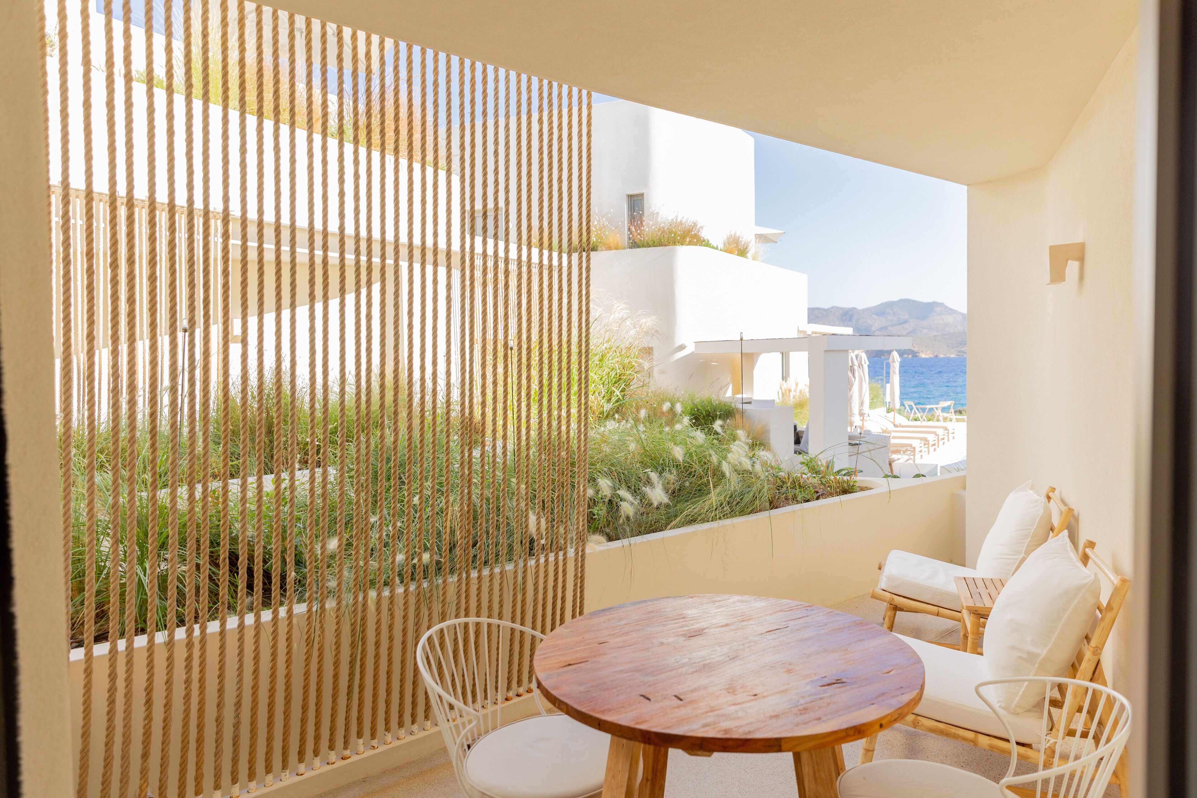 A balcony with a round wooden table and white wicker chairs, overlooking a coastal landscape with buildings and vegetation.