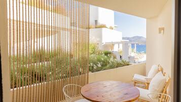 A balcony with a round wooden table and white wicker chairs, overlooking a coastal landscape with buildings and vegetation.