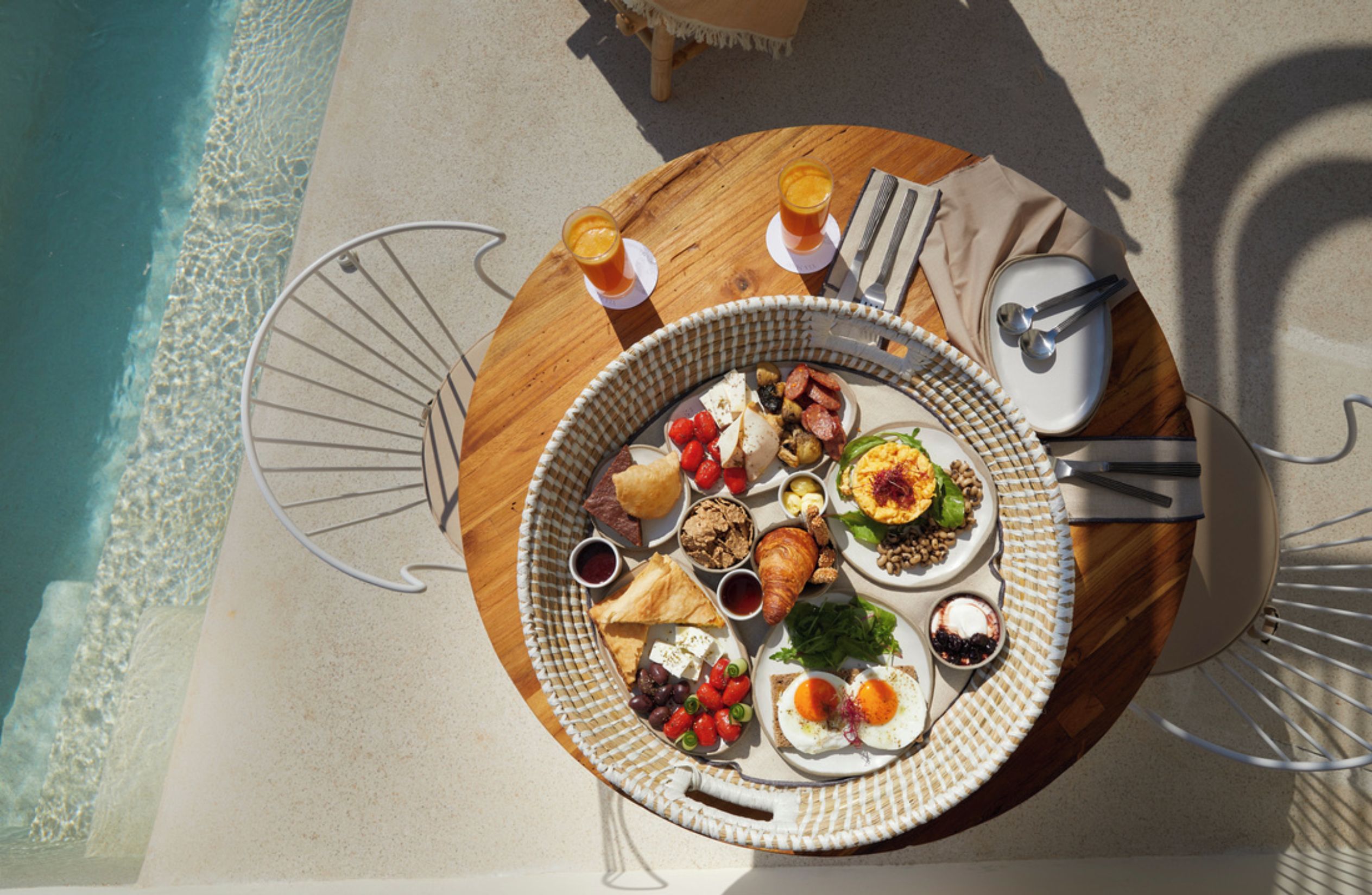 A round dining table with a variety of foods and drinks, set on a sandy beach with a pool in the background.