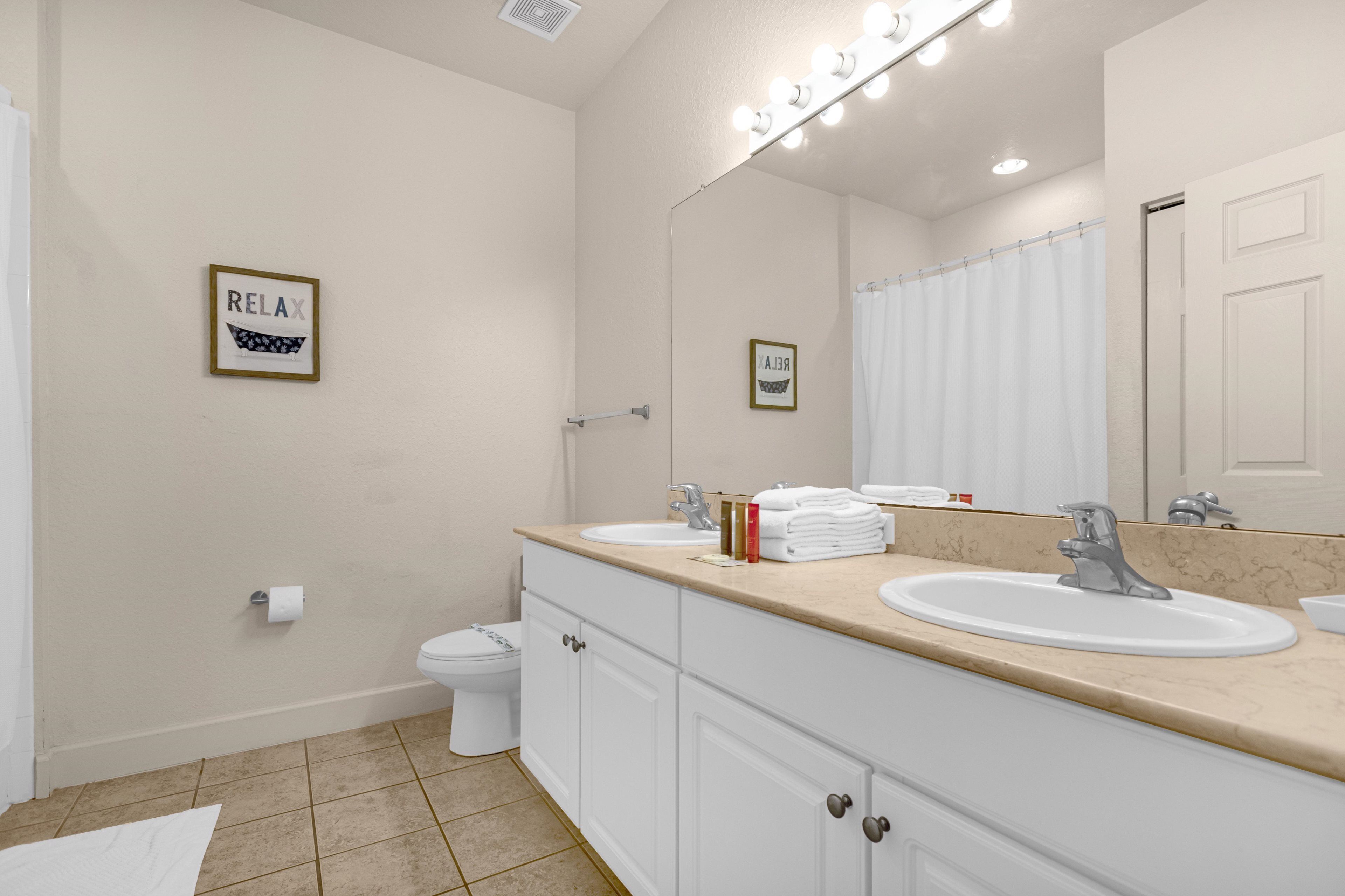 Une salle de bain avec deux lavabos, des toilettes, un rideau de douche et deux tableaux encadrés au mur.