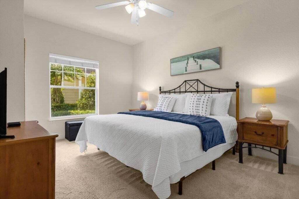 A bedroom with a bed, bedside lamps, a window, a ceiling fan, and a painting on the wall.