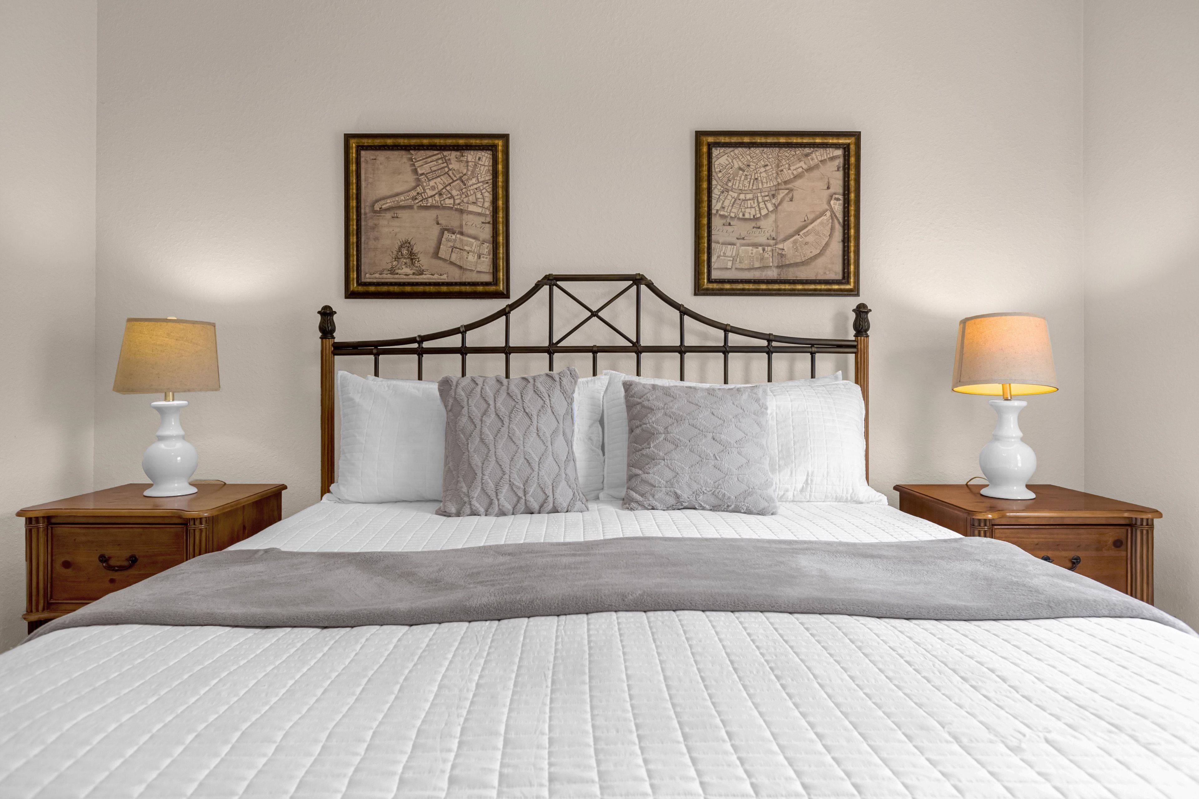 A double bed with a quilted white duvet, two nightstands with lamps, and two framed maps on the wall.
