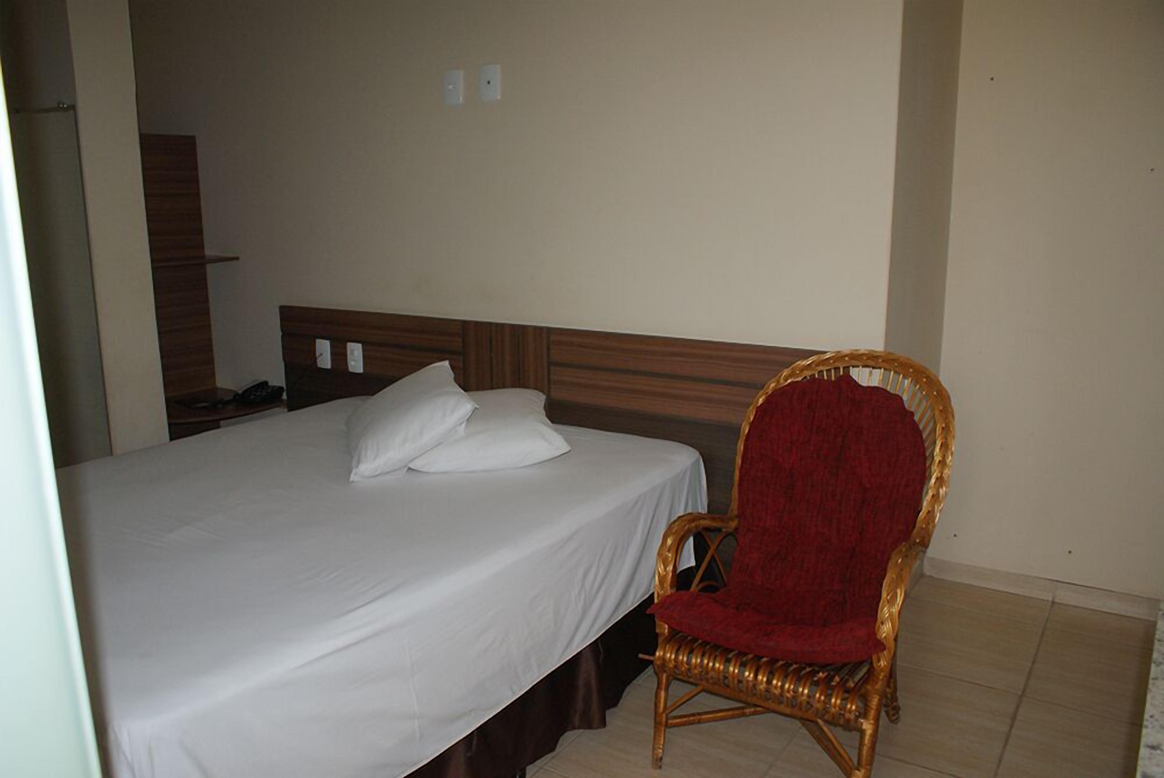 A single bed with white linens, a wooden headboard, and a wicker chair with a red cushion.