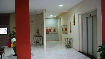Un vestíbulo de hotel con suelo de baldosas, un pilar rojo, una recepción, un reloj en la pared y un ascensor.