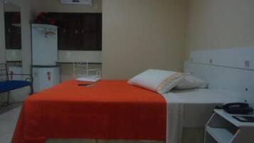 Una habitación pequeña con cama individual, cubierta roja, cabecero blanco y un escritorio pequeño con silla. Se observa un armario blanco y un espejo en la pared.