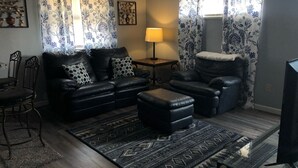 Una sala de estar con sofá, sillón, reposapiés y una mesa de centro de vidrio. Se observan dos lámparas, una alfombra y cortinas.