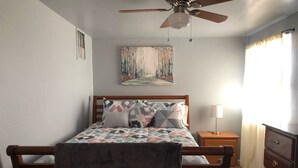 Un dormitorio con una cama de madera, un ventilador de techo, una lámpara de noche, un aparador y un cuadro en la pared.