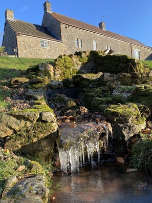 Un bâtiment en pierre, avec une formation rocheuse recouverte de mousse et un petit ruisseau qui en longe la base.