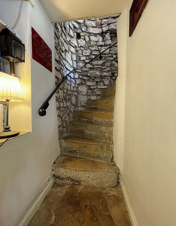 Un escalier étroit aux murs de pierre et agrémenté d’une main courante, menant à un palier où se trouvent une lampe et un panneau indicateur.