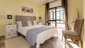 Un dormitorio con cama, mesitas de noche, una silla, ventana con cortinas y un cuadro en la pared.