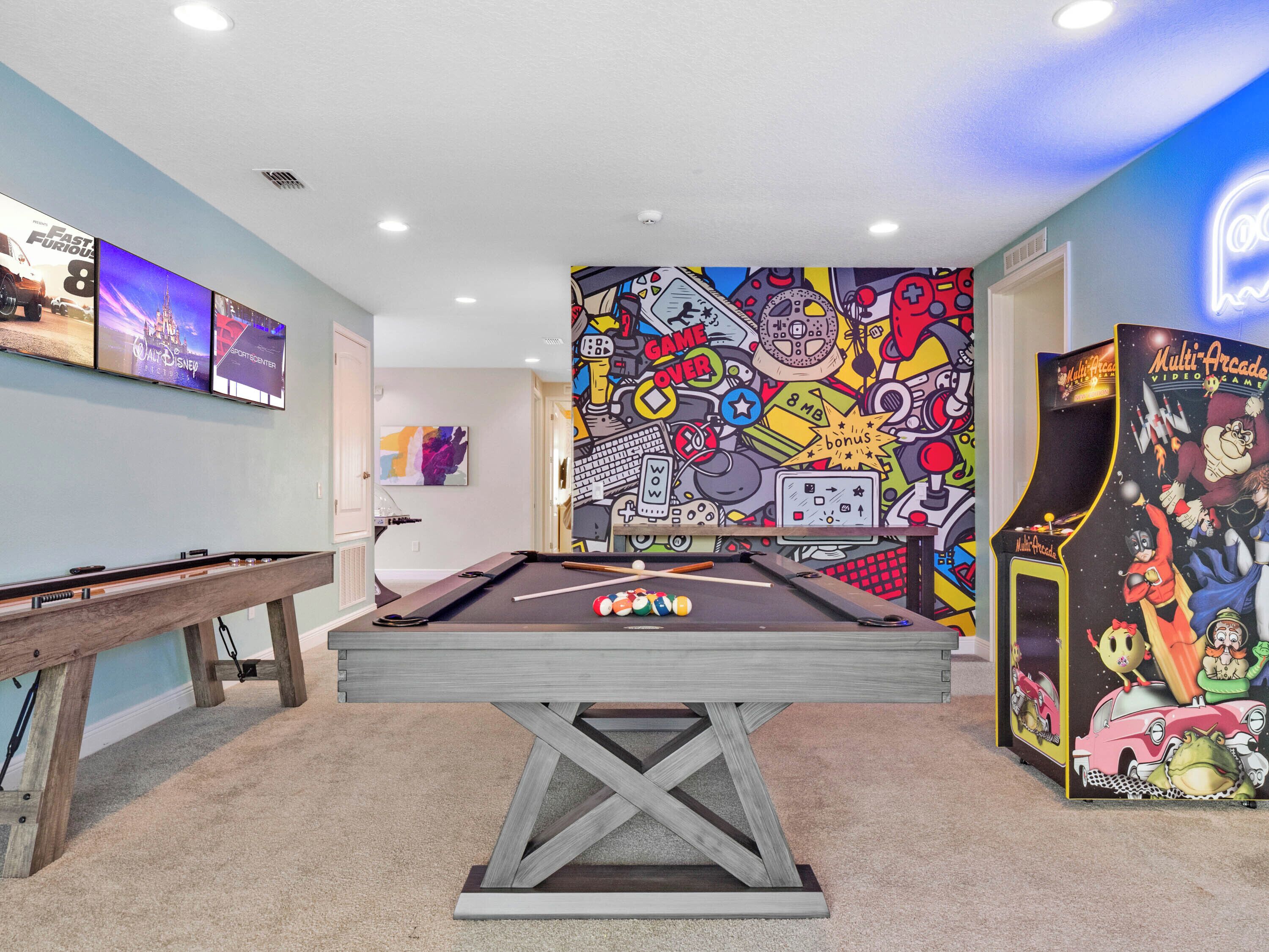 A pool table in a room with a colorful arcade-themed wall and a multi-arcade video game machine.
