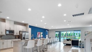 Cozinha moderna com armários brancos, uma ilha central com banquetas e uma área de jantar com uma grande janela com vista para um jardim.