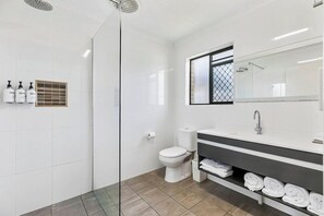 Ein modernes Badezimmer mit einer Glaskabine zur Dusche, einer weißen Toilette, einem Waschtisch mit großem Spiegel und ordentlich gefalteten Handtüchern auf einem Regal.