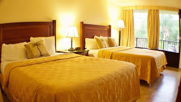 A hotel room with two beds, each with a wooden headboard, a nightstand with a lamp, and a window with curtains. - At Home Inn (Fort Pierce)