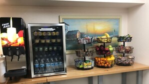 A refreshment station with a fruit juice dispenser, a refrigerator stocked with beverages, and a display of fruits and snacks on a wooden table. - At Home Inn (Fort Pierce)