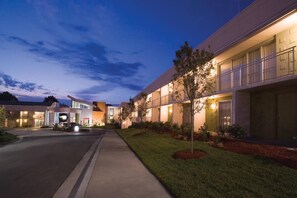 A hotel with a modern design, featuring a well-lit entrance, a covered walkway, and a landscaped area with trees.
