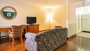 A room with a wooden desk, a television, a patterned sofa, a chair, and a lamp. - Rodeway Inn and Suites (Little Rock)