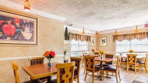A restaurant interior with wooden tables and chairs, a painting on the wall, and a window with blinds. - Rodeway Inn and Suites (Little Rock)