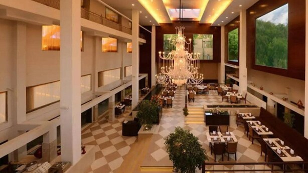 A spacious hotel lobby with multiple dining areas, a grand chandelier, and large windows offering views of greenery. - Jukaso IT Suites (Gurugram)