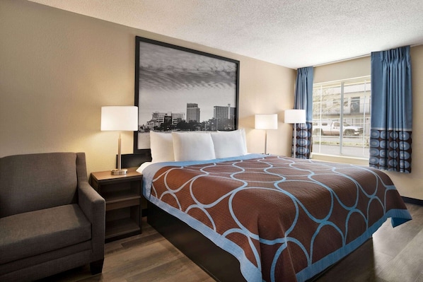 A hotel room with a bed, two bedside lamps, a chair, a large framed cityscape picture, and a window with curtains. - Super 8 By Wyndham Gulfport Near Biloxi (Gulfport)