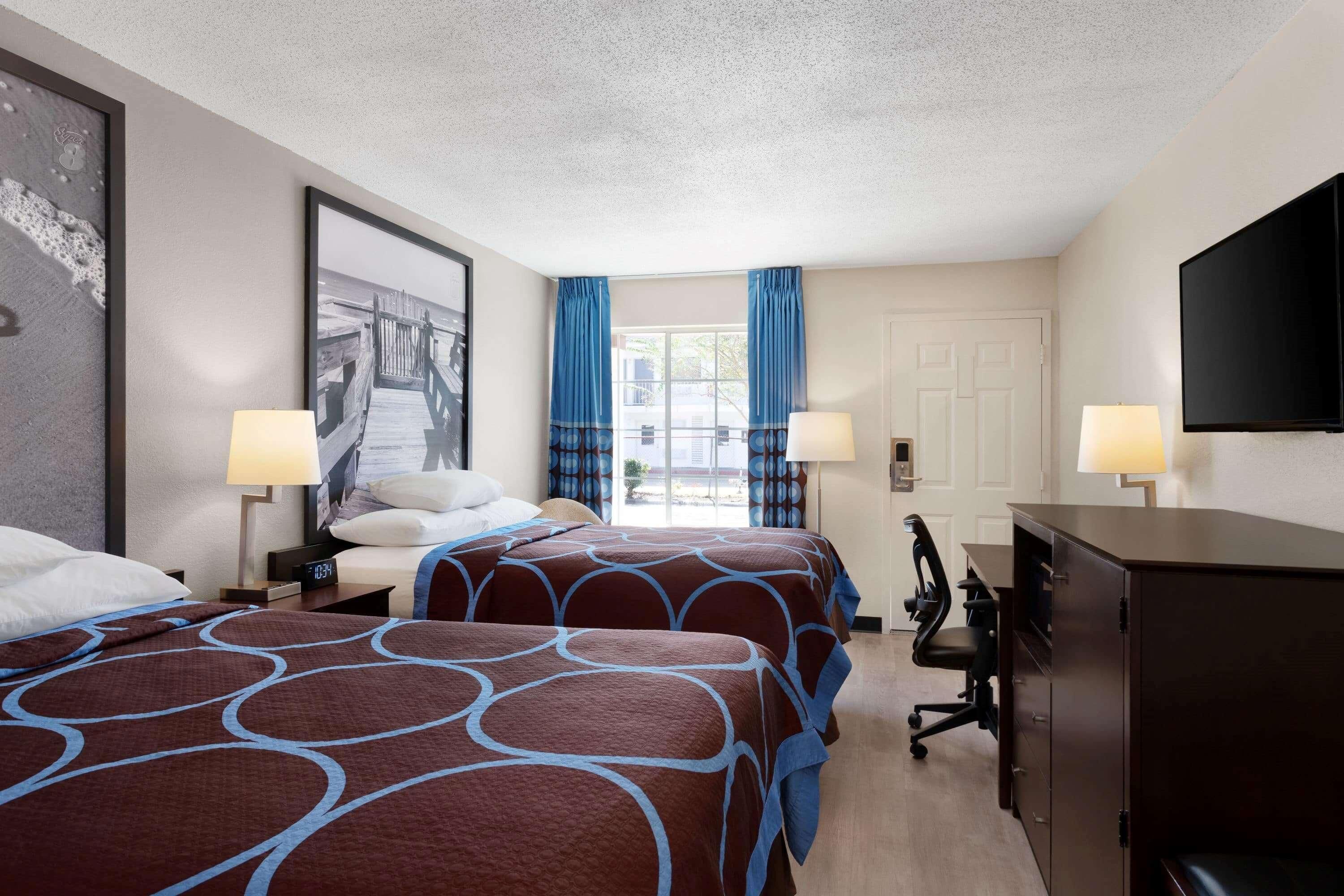 A hotel room with two beds, a desk, a chair, a TV, and a window with curtains.
