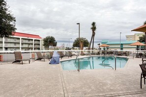 Una piscina con sillas de descanso y sombrillas, rodeada por una cerca blanca, con edificios al fondo.