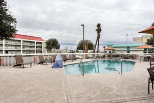 Una piscina con sillas de descanso y sombrillas, rodeada por una cerca blanca, con edificios al fondo.