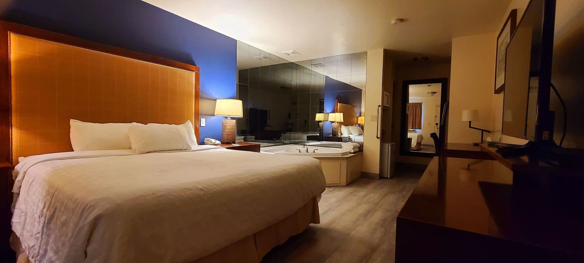 Habitación de hotel con una cama grande, un escritorio con silla, un espejo y un baño con lavamanos y espejo.