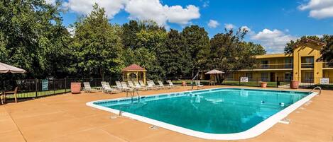 Une piscine rectangulaire, bordée d’une terrasse carrelée, entourée de chaises longues et de parasols, avec un kiosque et un bâtiment en arrière-plan.