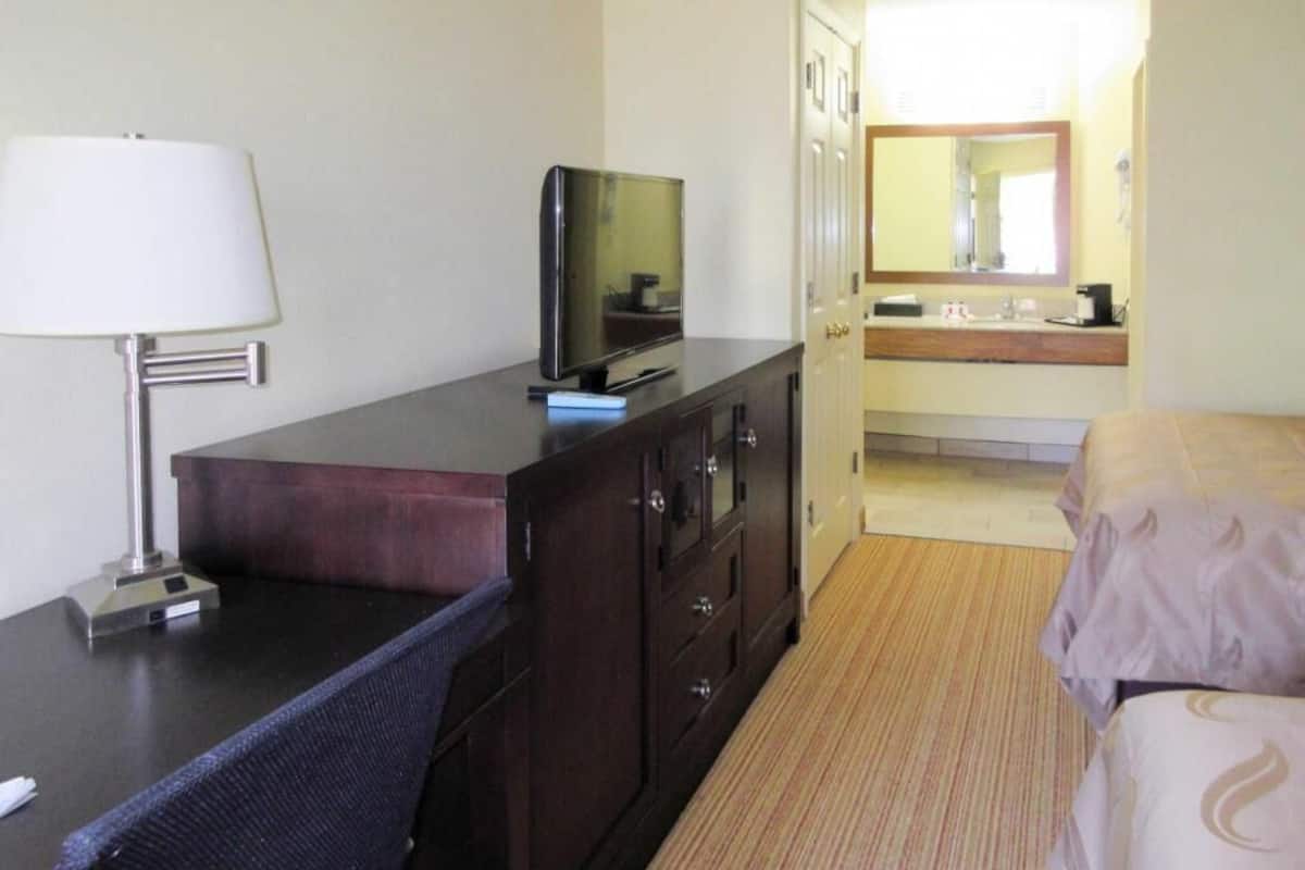A hotel room with a desk, a television, a bed, a bathroom with a mirror, and a carpeted floor.