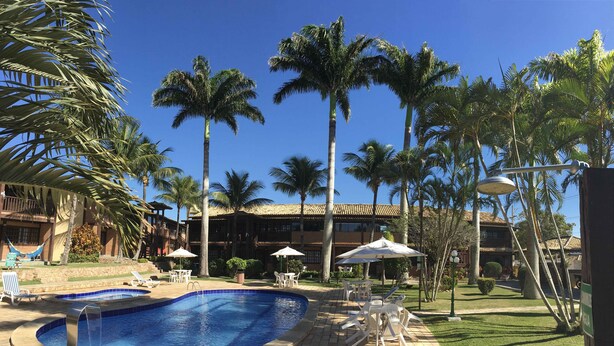 A pool area with lounge chairs and umbrellas, surrounded by palm trees and a building with a tiled roof. - Cozy property just a 3-minute walk from the beach (Free Wifi Room Service) (Buzios)