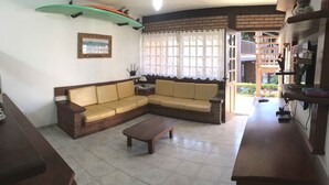 A living room with a sofa, a coffee table, and a TV. There is a window with curtains and a view of the outside. - Cozy property just a 3-minute walk from the beach (Free Wifi Room Service) (Buzios)