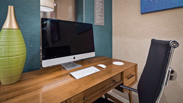 A wooden desk with an Apple iMac, a keyboard, and a mouse. There is a green vase on the left and a black chair to the right.